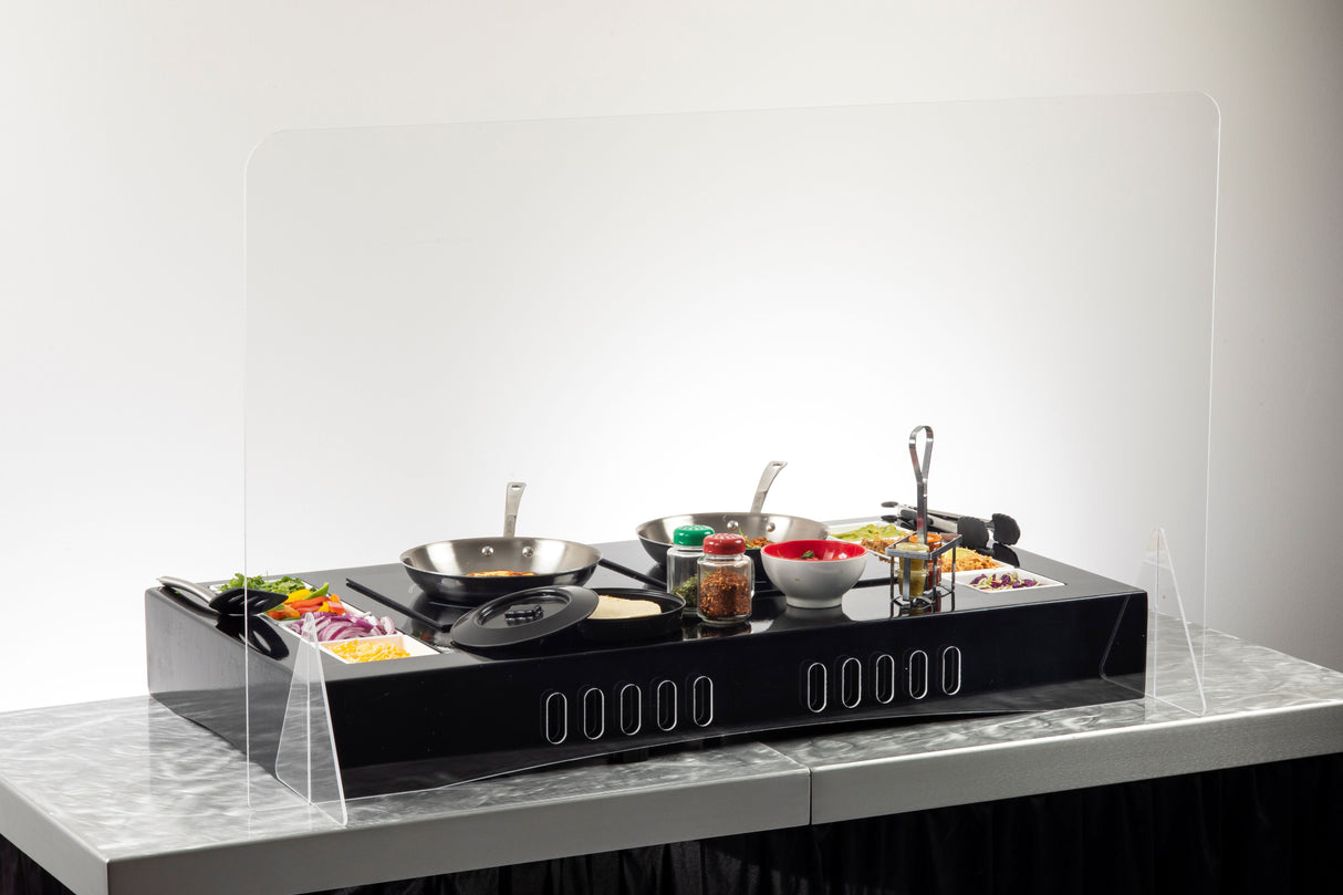 A black tabletop cooking station with two frying pans, bowls of ingredients, utensils, and seasonings is set behind a clear acrylic shield on a marble-like surface with a white background.