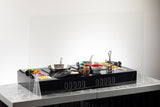 A black tabletop cooking station with two frying pans, bowls of ingredients, utensils, and seasonings is set behind a clear acrylic shield on a marble-like surface with a white background.