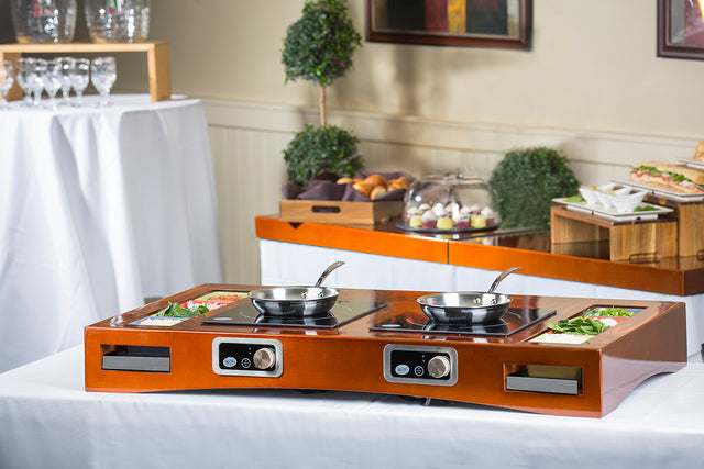 A polished wood buffet features a Double induction countertop station with two burners, each holding a frying pan. Cut vegetables rest in built-in compartments. Behind, pastries and sandwiches sit under glass domes amid elegant greenery and glassware.