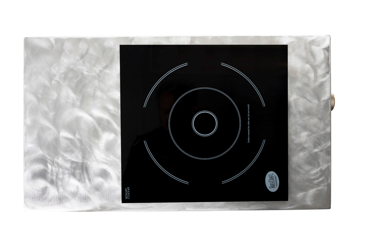 A rectangular, silvery textured metal surface with a shiny finish features a TableCraft Products Induction Station, showcasing a black glass single burner cooktop with white circular outlines, touch controls, and a round badge labeled INDUCTION.