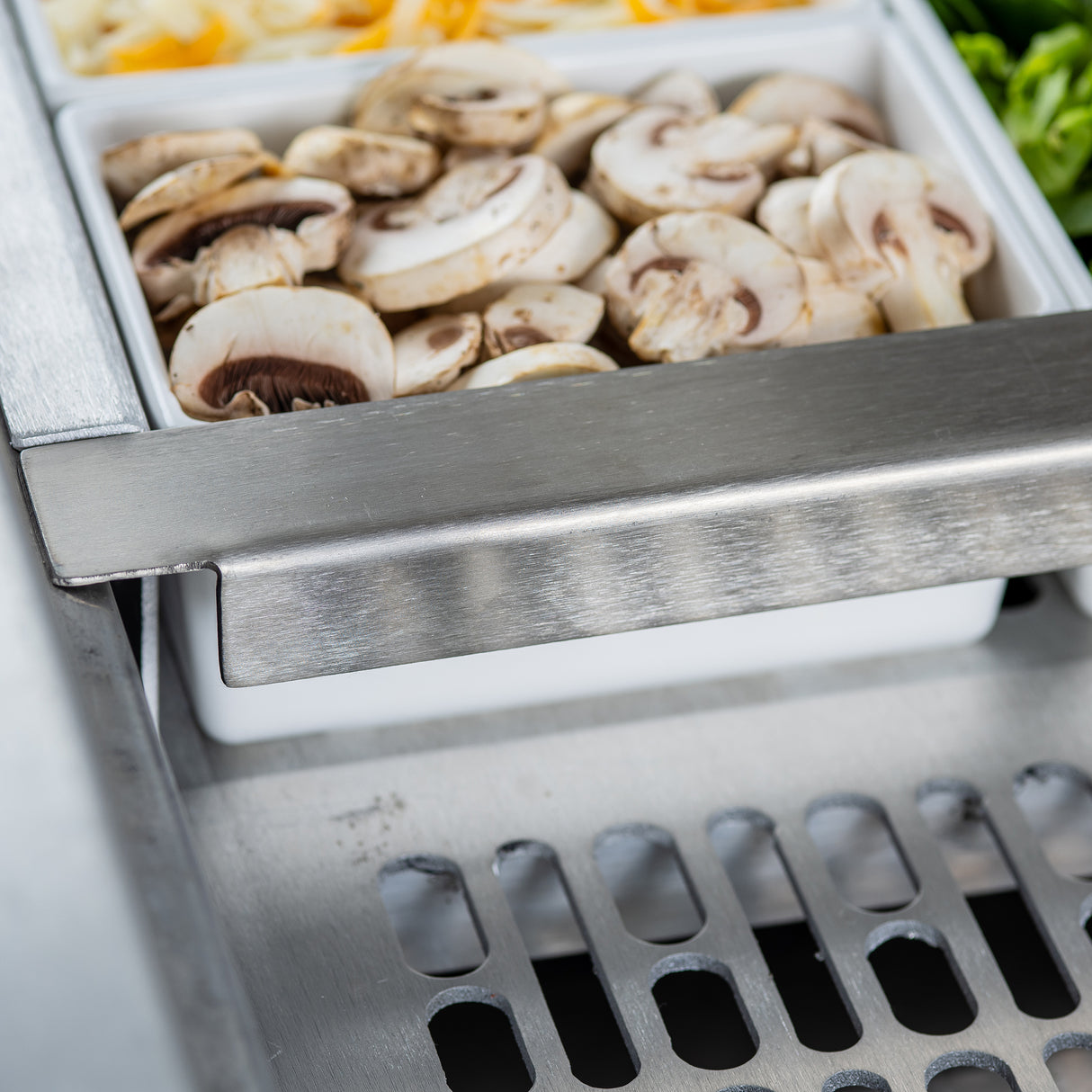 Close-up of a stainless steel salad bar with a white container filled with sliced fresh mushrooms. In the background are shredded cheese and leafy greens, set in compartments. The shiny metal surface and ventilation grate by TableCraft Products are visible in front.