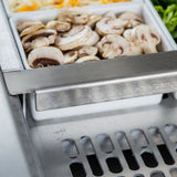 Close-up of a stainless steel salad bar with a white container filled with sliced fresh mushrooms. In the background are shredded cheese and leafy greens, set in compartments. The shiny metal surface and ventilation grate by TableCraft Products are visible in front.