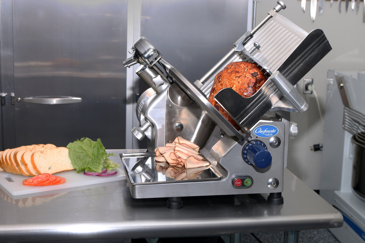 A Globe GC512 commercial meat slicer sits on a stainless steel counter, slicing roasted meat into thin layers. Nearby are sliced bread, lettuce, tomato, and onion on a cutting board, with kitchen utensils and a metal cabinet in the background.