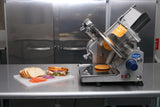 A Globe GC512 commercial deli slicer sits on a stainless steel counter with a block of cheese loaded inside. In front, a cutting board displays sliced bread, tomatoes, onions, and a made sandwich. Silver cabinets are visible in the background.