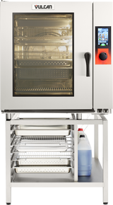 A Vulcan TCM-102E electric steam oven with a glass door reveals multiple wire racks. To the right, a digital control panel features a display and red knob. Below, a metal stand holds trays and a large container of cleaning solution.