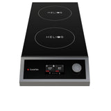 A black and silver double induction cooktop with a portable design features two circular burners labeled “HELIOS.” The front panel offers a digital display, single control knob, CookTek logo, and a red indicator light.
