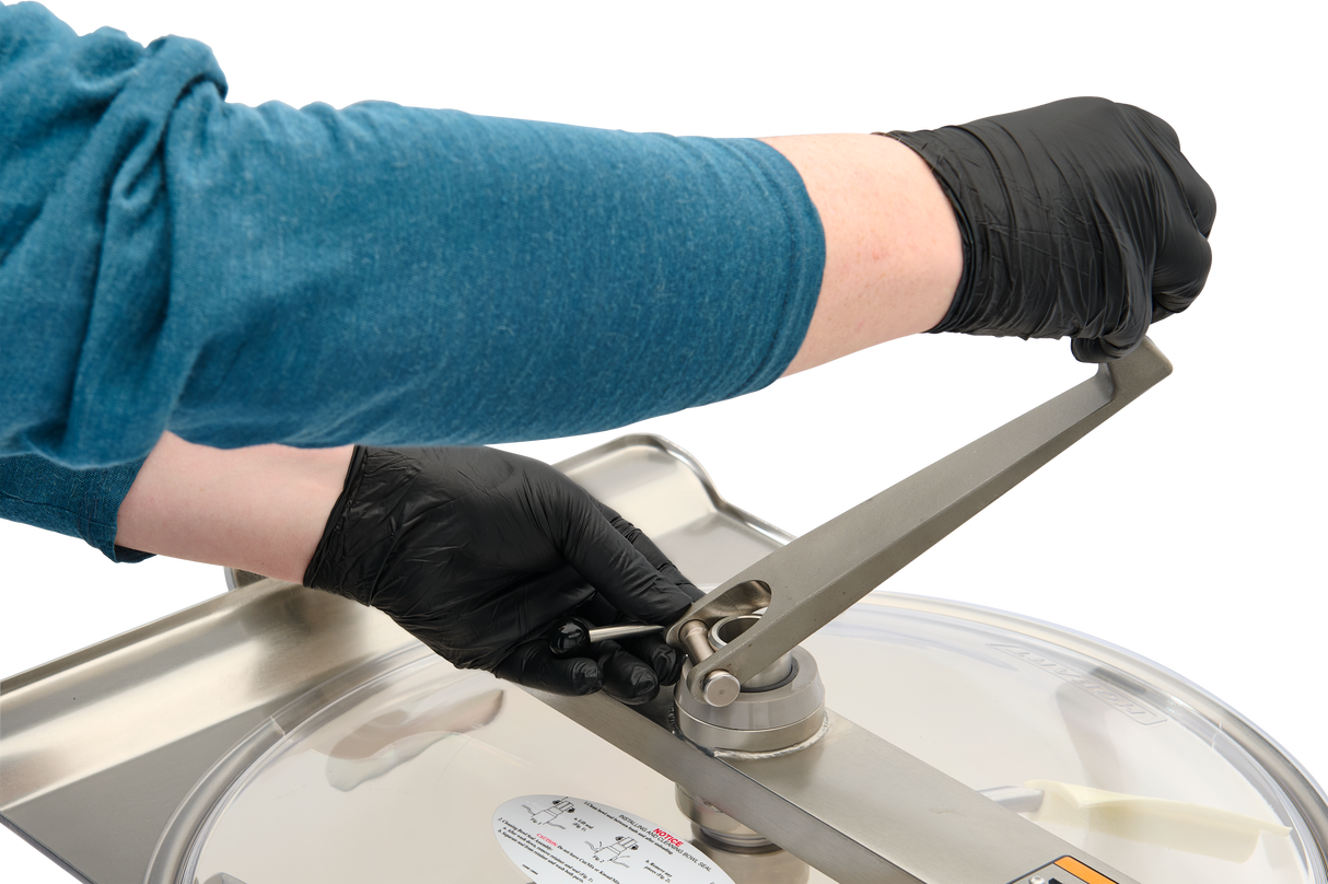A person wearing black gloves and a teal long-sleeve shirt uses a metal tool to adjust a bolt on a Hobart Cutter Mixer with a stainless steel bowl and clear lid, focusing on the hand and hardware details. The background is plain and light.