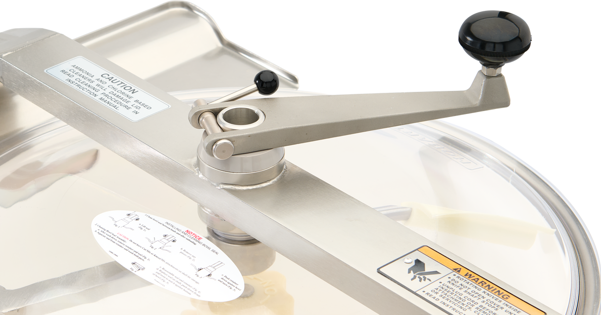 A close-up of a metal crank handle with a black knob, attached to a Hobart HCM450C-61 cutter mixer lid. The stainless steel lid has caution labels, a transparent window, and visible screws for attachment. The background is light and neutral.