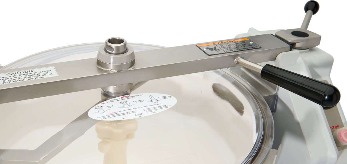 Close-up of a Hobart Cutter Mixer autoclave lid with a black handle, warning labels, instruction sticker, and a round transparent section at the center. The plain white background highlights its safety features and stainless steel bowl design.