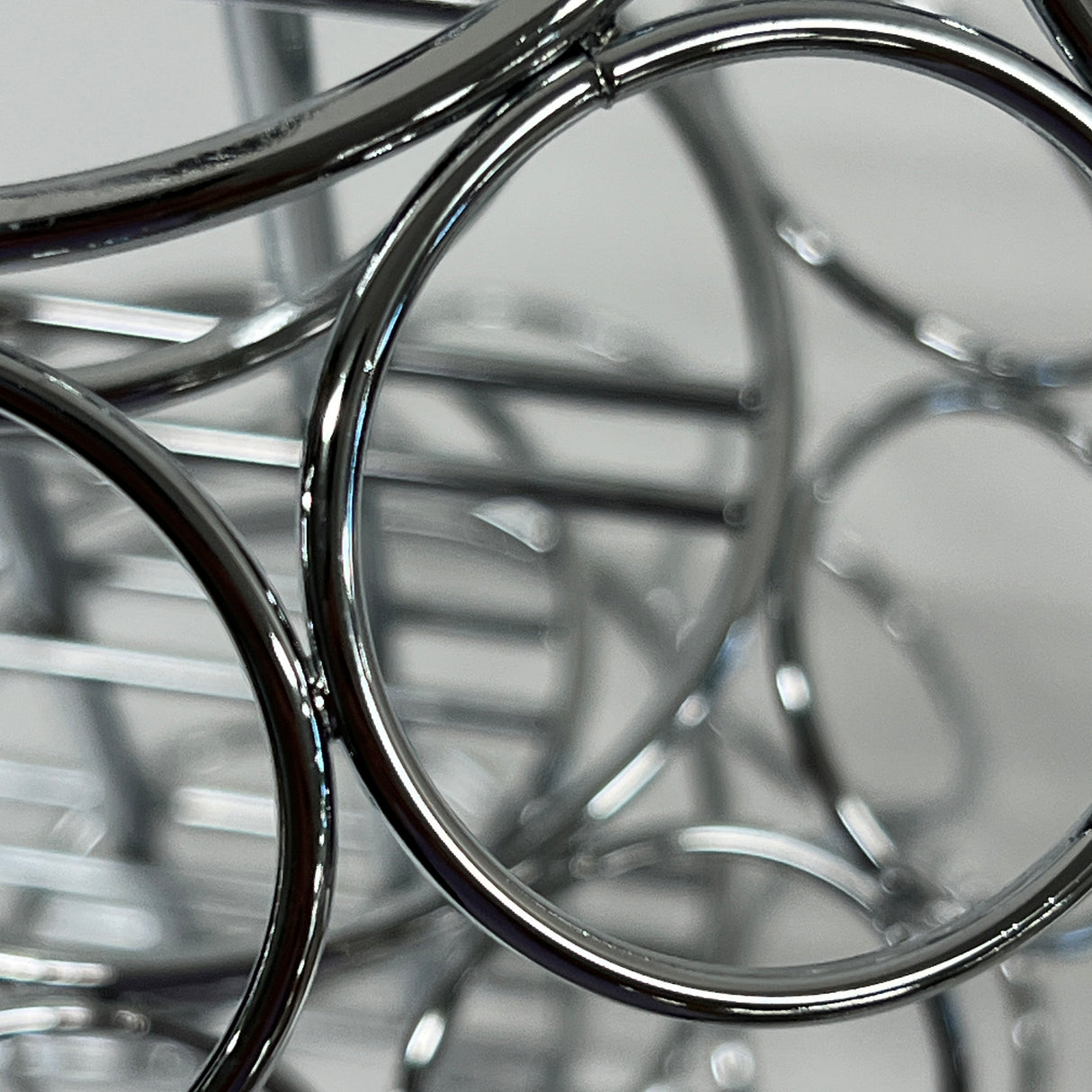 Close-up photo of shiny, overlapping metal rings and bars from a TableCraft 3-tier cone holder, creating an abstract pattern with reflections. The softly blurred background highlights the polished, chrome-like surfaces and curved shapes in the foreground.