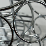 Close-up photo of shiny, overlapping metal rings and bars from a TableCraft 3-tier cone holder, creating an abstract pattern with reflections. The softly blurred background highlights the polished, chrome-like surfaces and curved shapes in the foreground.