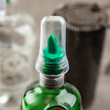 A close-up of a green glass bottle with a BPA free plastic liquor pourer cover and clear cap. The pour spout is green. The bottle reads “London Dry.” Blurred items, like another bottle and a dark cylindrical container, are in the background.