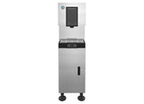 A stainless steel icemaker dispenser stands upright on four black legs. The top section features a black dispensing area with push levers, a drip tray below, and a blue logo in the top left corner. The lower section is plain with a narrow vertical window.