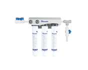 A wall-mounted Blueair triple filtration system with three vertical filter cartridges, labeled with instructions. Ideal for ice machine filtration and scale reduction, the unit features two pressure gauges and blue valve controls on white piping.