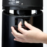 A hand presses a lever on a black cylindrical beverage dispenser, likely an insulated hot water appliance. The label above shows “COLOR: BLACK” and other product details in white text against a plain white background.