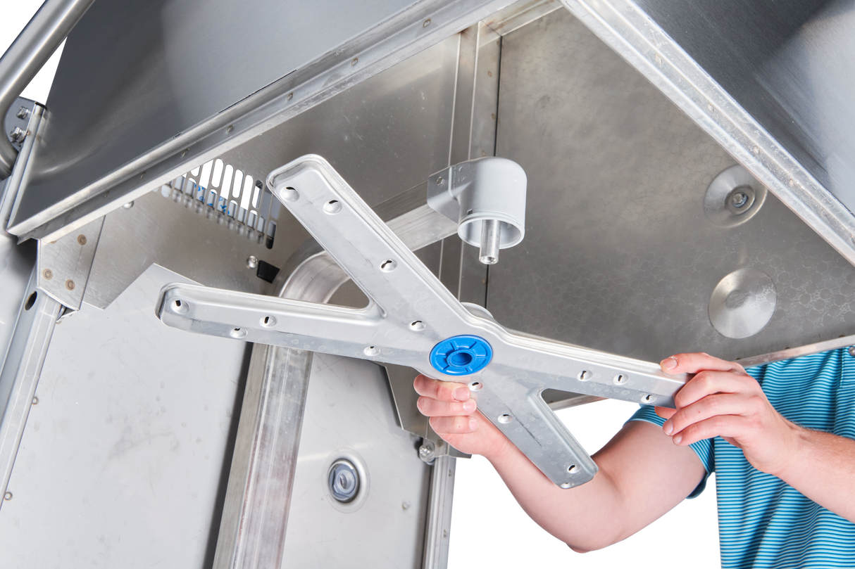 A person in a blue striped shirt installs or removes a metal spray arm inside a commercial dishwasher, with their hands gripping the arm and the open door revealing the stainless steel interior and various components.