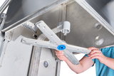 A person in a blue striped shirt installs or removes a metal spray arm inside a commercial dishwasher, with their hands gripping the arm and the open door revealing the stainless steel interior and various components.