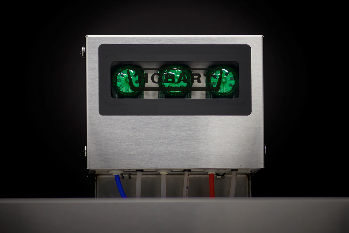 A stainless steel commercial dishwasher features three green illuminated indicator lights behind a dark window labeled HOBART. Tubes colored blue, clear, and red emerge from the bottom, and the dark background highlights this chemical sanitizing equipment.