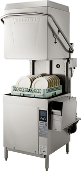 A commercial stainless steel ventless dishwashing machine with a pull-down hood, labeled Hobart. Inside, a green rack holds clean white plates. Control panel and soap dispenser are visible, with pipes and wiring at the side and back.