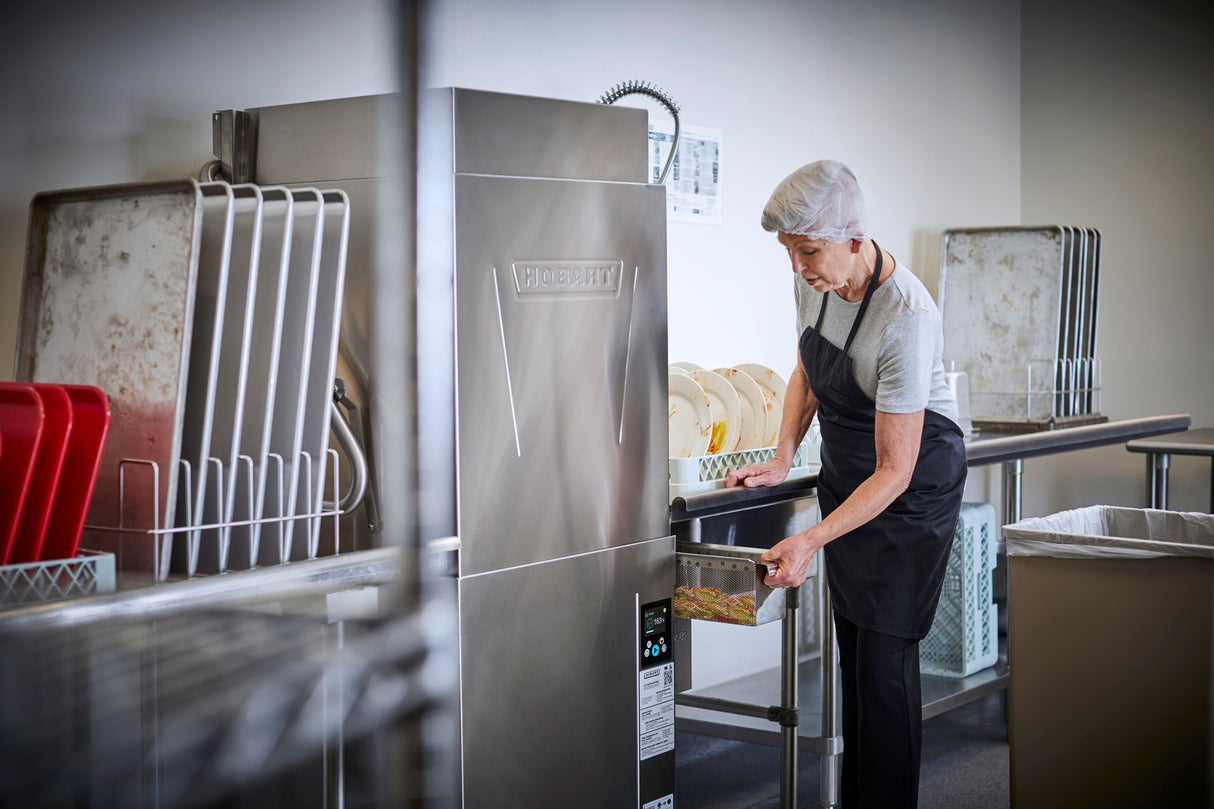 An older woman in a hairnet, gray shirt, and black apron loads dishes into a high temp sanitizing ventless dishwashing machine. Clean dishes are stacked on metal racks nearby. The kitchen is clean and organized, with a trash bin visible beside her.