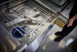 A close-up of a high temp sanitizing, ventless dishwashing machine shows an open interior with metal racks and spray arms. A person’s hand pulls out a mesh tray filled with food debris. The metallic space gleams with blue accents, while the grey floor is speckled.