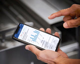 A person holds a smartphone in one hand and uses the other to interact with a screen displaying a bar graph and financial data in blue and gray, possibly analyzing energy recovery trends. The blurred background draws focus to the hands and phone.