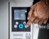 A hand presses a button on a sleek, modern Hobart ventless dishwashing machine control panel. The display shows a high temp sanitizing rinse at 183°F, with water and play icons, labeled buttons, and a QR code below the screen.