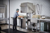 A person wearing a hairnet and uniform loads dishes into a ventless dishwashing machine in a commercial kitchen. Stacks of plates sit on metal racks, while white tiled walls and stainless steel surfaces emphasize the clean, organized environment.