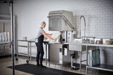 A person wearing a hairnet, gray shirt, and black apron loads dishes into a large ventless dishwashing machine in a clean, tiled kitchen. Racks of plates and utensils line the stainless steel counters on either side of the high temp sanitizing machine.