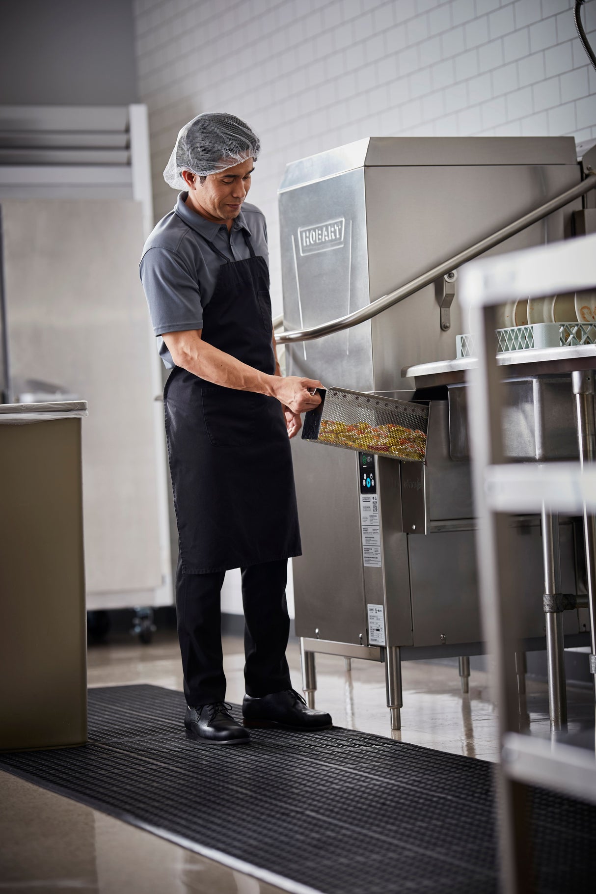 A man wearing a hairnet, black apron, polo shirt, and black pants loads a tray with dirty dishes into a ventless dishwashing machine in a commercial kitchen with white tile walls and stainless steel equipment.