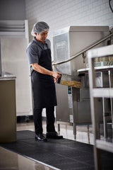 A man wearing a hairnet, black apron, polo shirt, and black pants loads a tray with dirty dishes into a ventless dishwashing machine in a commercial kitchen with white tile walls and stainless steel equipment.
