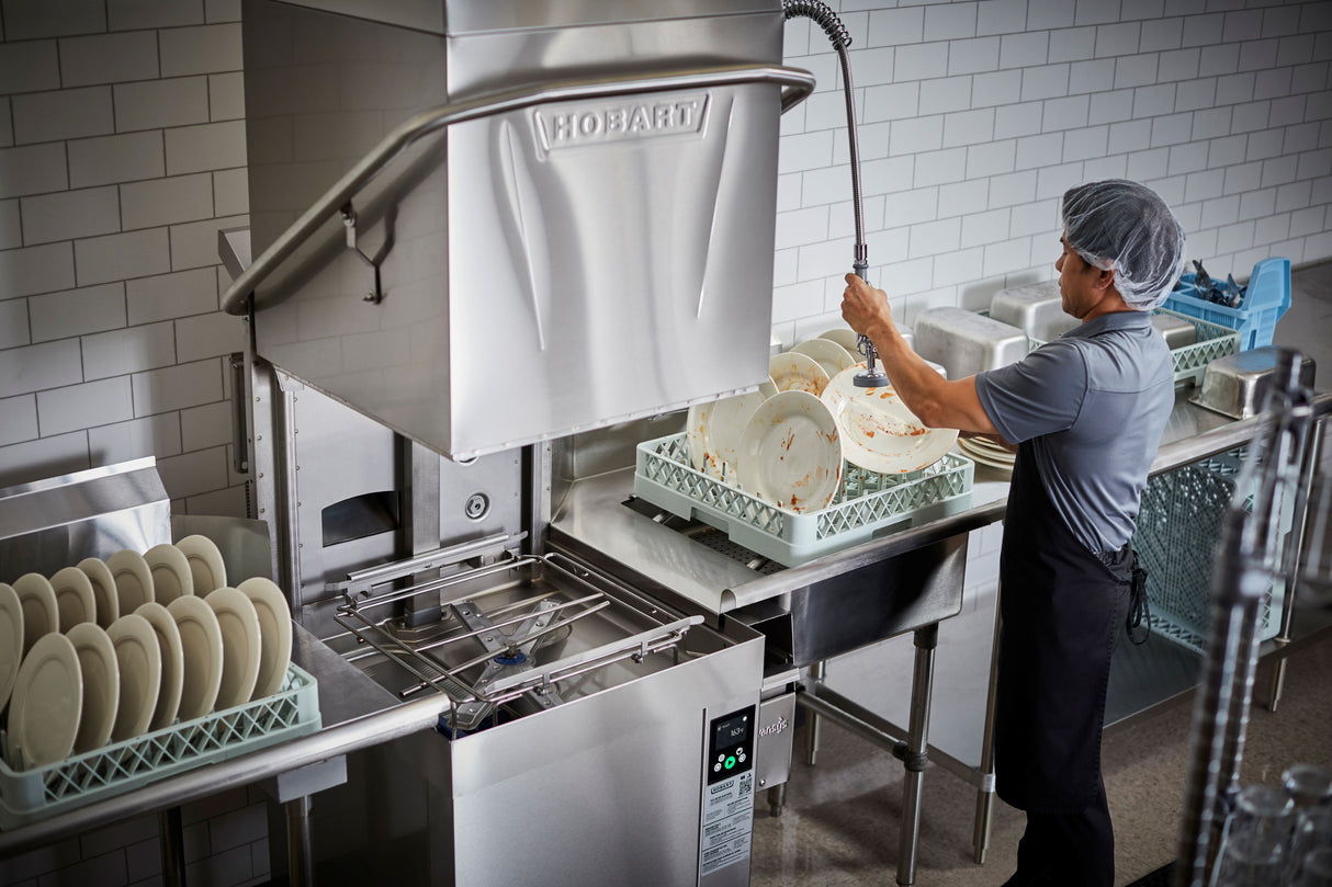 A person in a hairnet, apron, and gray shirt sprays water onto dirty plates in a bright commercial kitchen with a ventless dishwashing machine. Clean plates are stacked nearby as the high temp sanitizing cycle keeps the kitchen orderly and efficient.