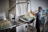 A person in a hairnet, apron, and gray shirt sprays water onto dirty plates in a bright commercial kitchen with a ventless dishwashing machine. Clean plates are stacked nearby as the high temp sanitizing cycle keeps the kitchen orderly and efficient.