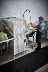 A person in a gray shirt, black apron, and hairnet uses a Hobart ventless dishwashing machine. They spray water on dishes stacked in racks beside the machine in a clean kitchen with white tiled walls and black floor mats.