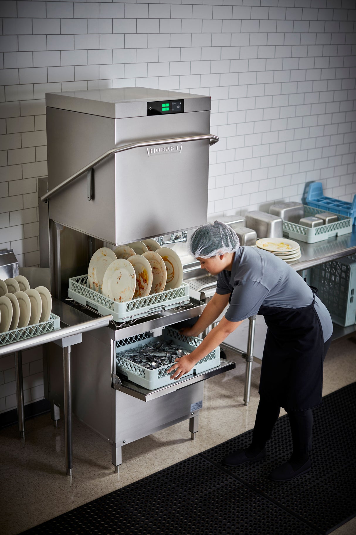 A worker wearing a hairnet and uniform loads dirty dishes into a Two Level Door Type Dishwasher in a clean, tiled kitchen. The high temp sanitizing unit handles up to 80 racks/hr, with organized plates and utensils and stacks of clean dishes nearby.