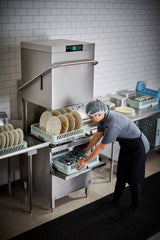 A worker wearing a hairnet and uniform loads dirty dishes into a Two Level Door Type Dishwasher in a clean, tiled kitchen. The high temp sanitizing unit handles up to 80 racks/hr, with organized plates and utensils and stacks of clean dishes nearby.
