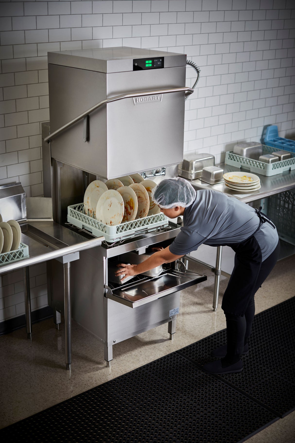 A person in a hairnet, gray shirt, and black pants loads dirty dishes into a Two Level Door Type Dishwasher with high temp sanitizing, capable of handling up to 80 racks/hr in a kitchen with white tiled walls and stainless steel counters.