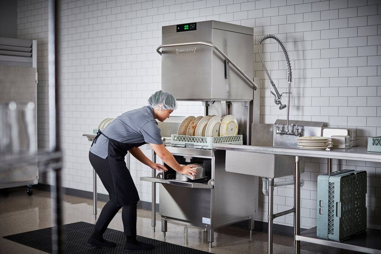 A person wearing a hairnet, gloves, and uniform loads plates into a Two Level Door Type Dishwasher in a commercial kitchen. The 80 racks/hr high temp sanitizing machine keeps the white-tiled space spotless and efficient.
