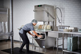 A person wearing a hairnet, gloves, and uniform loads plates into a Two Level Door Type Dishwasher in a commercial kitchen. The 80 racks/hr high temp sanitizing machine keeps the white-tiled space spotless and efficient.