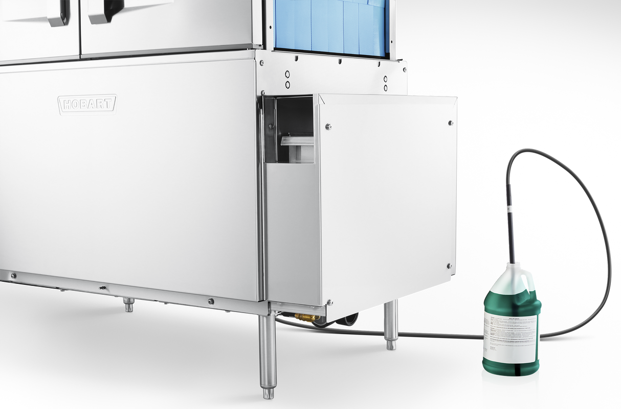 A stainless steel commercial conveyor dishwasher with a lower compartment open, revealing an internal container. A black hose connects the machine to a green and white detergent jug, highlighting automatic soil removal, against a black background.