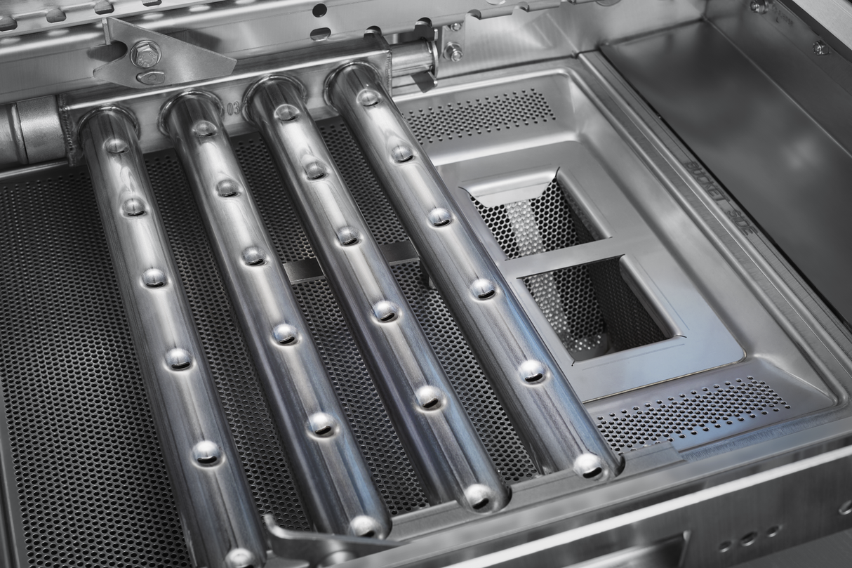 Close-up of the interior of a stainless steel gas grill, showing four metal burner tubes with small holes for flame and two rectangular heat shields. The clean surfaces shine, similar to those maintained by automatic soil removal in commercial appliances.