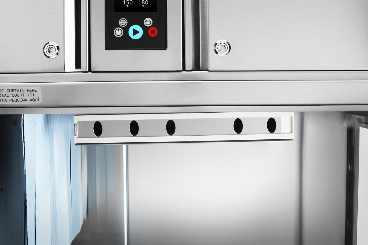 Close-up of a stainless steel conveyor dishwasher with a control panel featuring a display and buttons. Below, horizontal vent openings hint at automatic soil removal, while the blue curtain and reflective interior are visible on either side.