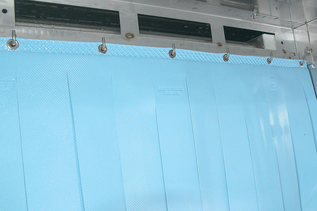 A close-up of a light blue plastic strip curtain hanging from metal hooks on a stainless steel frame, with vertical ridges. The background features reflective metallic surfaces, typical of an ENERGY STAR Hobart FT1000E low energy flight type dish machine.
