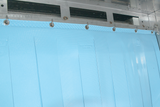A close-up view of a light blue, flexible plastic sheet hanging from metal hooks in a ventless flight type dish machine. The sheet features a textured pattern and embossed markings, attached to an industrial fixture with an automatic soil removal system.