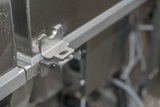 Close-up of a metal bracket attached to the corner of a stainless steel Hobart FT1000E flight type dish machine. The bracket is secured by a hinge, and a grey rubber seal runs along the edge. Wires and components are visible in the background.