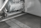 Close-up of an industrial metallic machine part featuring a cylindrical electric motor with ventilation holes, metal tubing, and a perforated floor grid—details common to a Hobart FT1000E flight type dish machine in a gray, utilitarian setting.