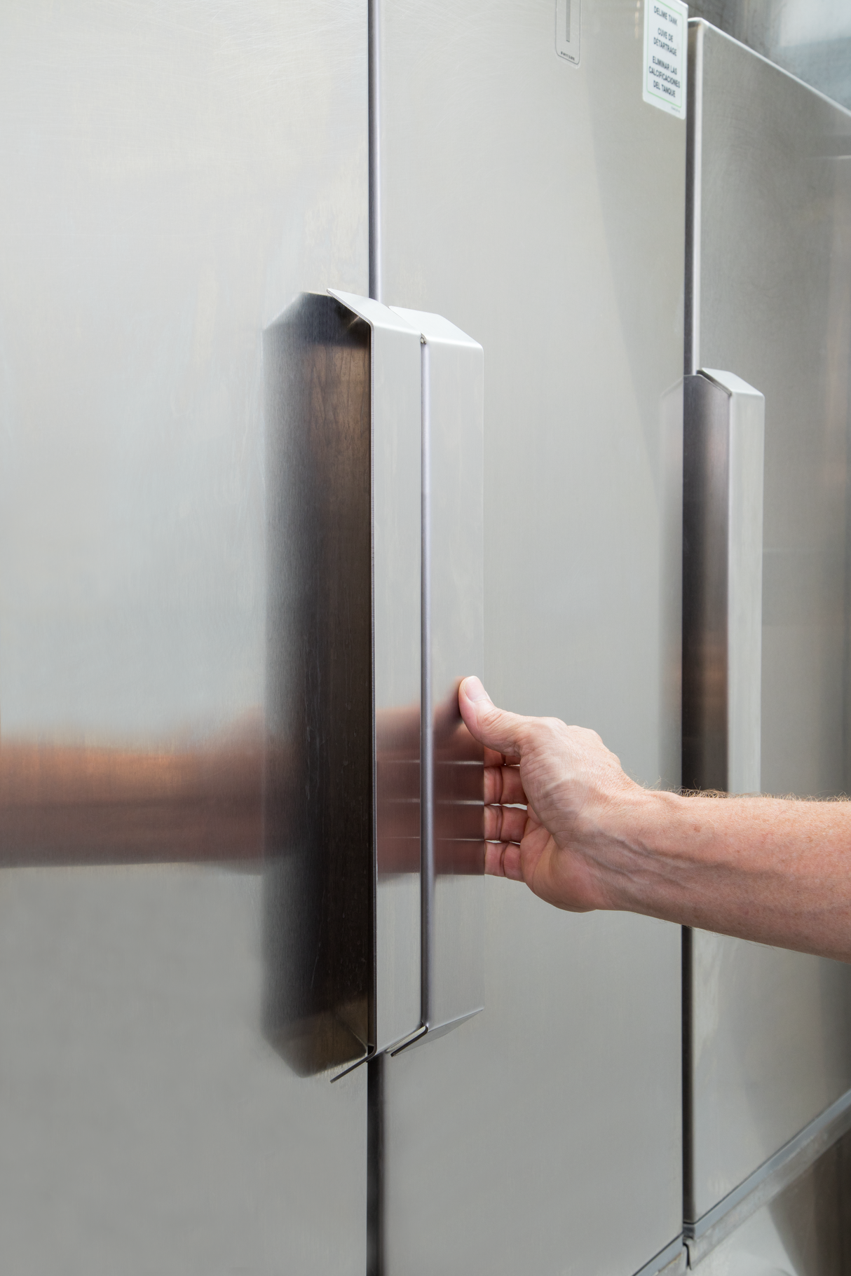 A person’s right hand grips the vertical metal handle of a large stainless steel commercial refrigerator door. The surface reflects light, and another similar handle—like those found on low energy series equipment—is visible in the background.