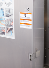Close-up of a stainless steel flight type dish machine door with a metal latch. A white warning label with orange stripes and text is affixed next to blue and white illustrated step-by-step instructions from the low energy series.