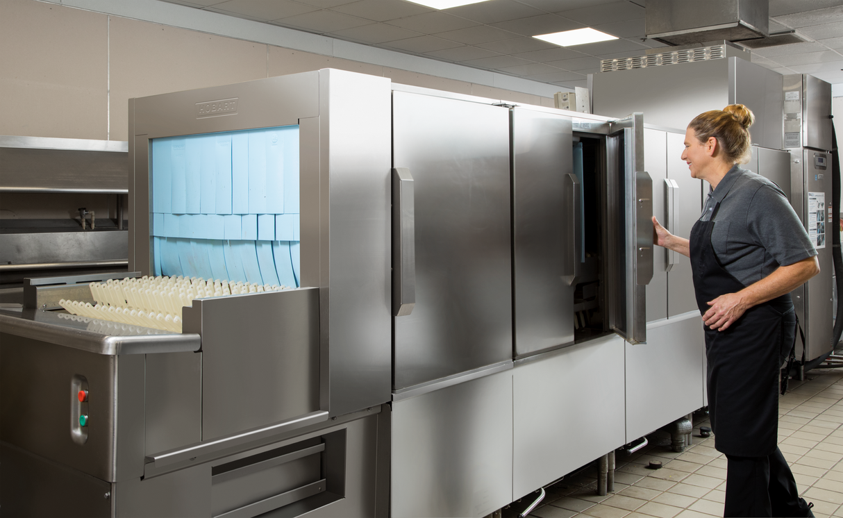 A woman in a black apron and gray shirt operates a flight type dish machine in a commercial kitchen with stainless steel equipment. Clean trays and utensils are stacked neatly, while the energy recovery system helps maintain efficiency in the brightly lit space.