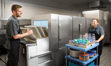Two people in a commercial kitchen, both in gray uniforms, load and unload a Hobart FT1000E flight type dish machine. One manages trays at the low energy dish machine, while the other moves a cart with clean dishes through the stainless steel workspace.
